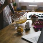 A woman uses an electric mixer to whip eggs in a cozy kitchen setting, surrounded by fruits and a laptop.